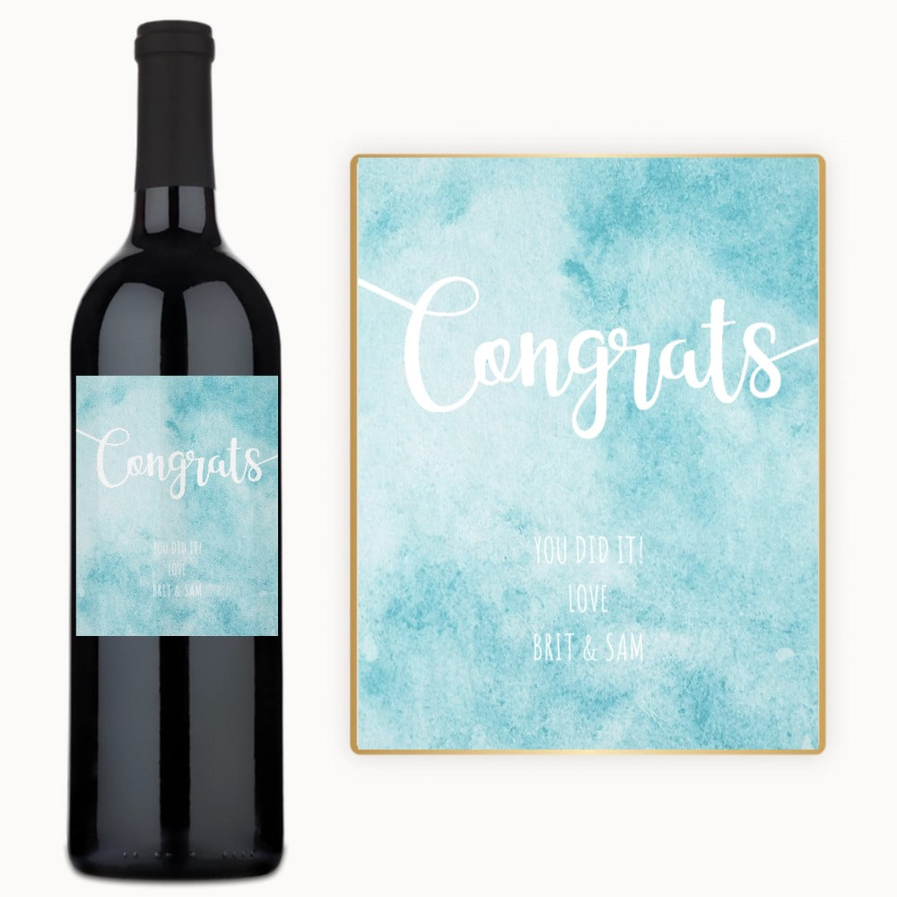 A bottle of wine with a custom label next to a blue watercolor Congrats card.