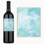 A bottle of wine with a custom label next to a blue watercolor Congrats card.