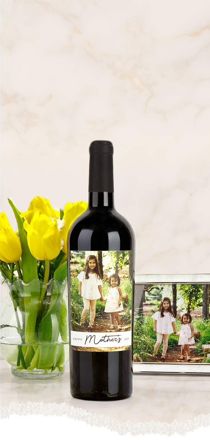 Red wine bottle with a custom label featuring a family photo, next to a vase of yellow tulips and a framed picture.