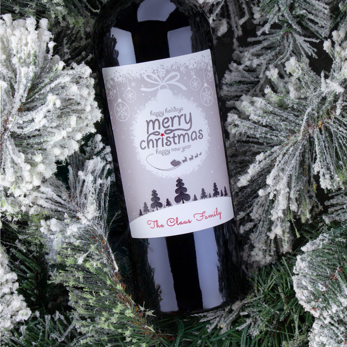 A personalized wine bottle labeled The Clear Family sits among snowy pine branches as a Christmas ornament.