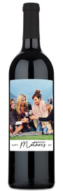 Three women smiling outdoors, holding a wine bottle with a custom Happy Mothers Day label.