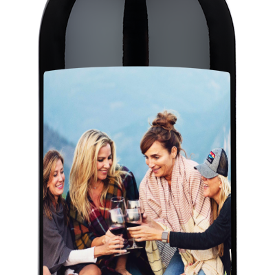 Three women smiling outdoors, holding a wine bottle with a custom Happy Mothers Day label.