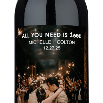 A couple kisses at their wedding surrounded by guests holding sparklers; a personalized wine bottle is in the foreground.