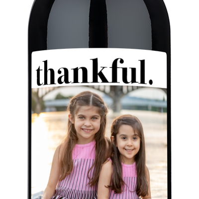 Two smiling girls outdoors on a wine bottle label with thankful in script and a holiday photo.