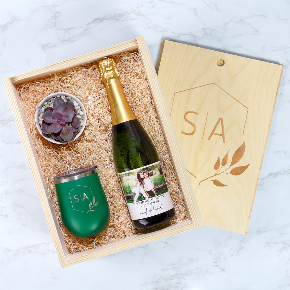 A gift box with a small succulent, green tumbler, and two champagne bottles in a double wood wine box.